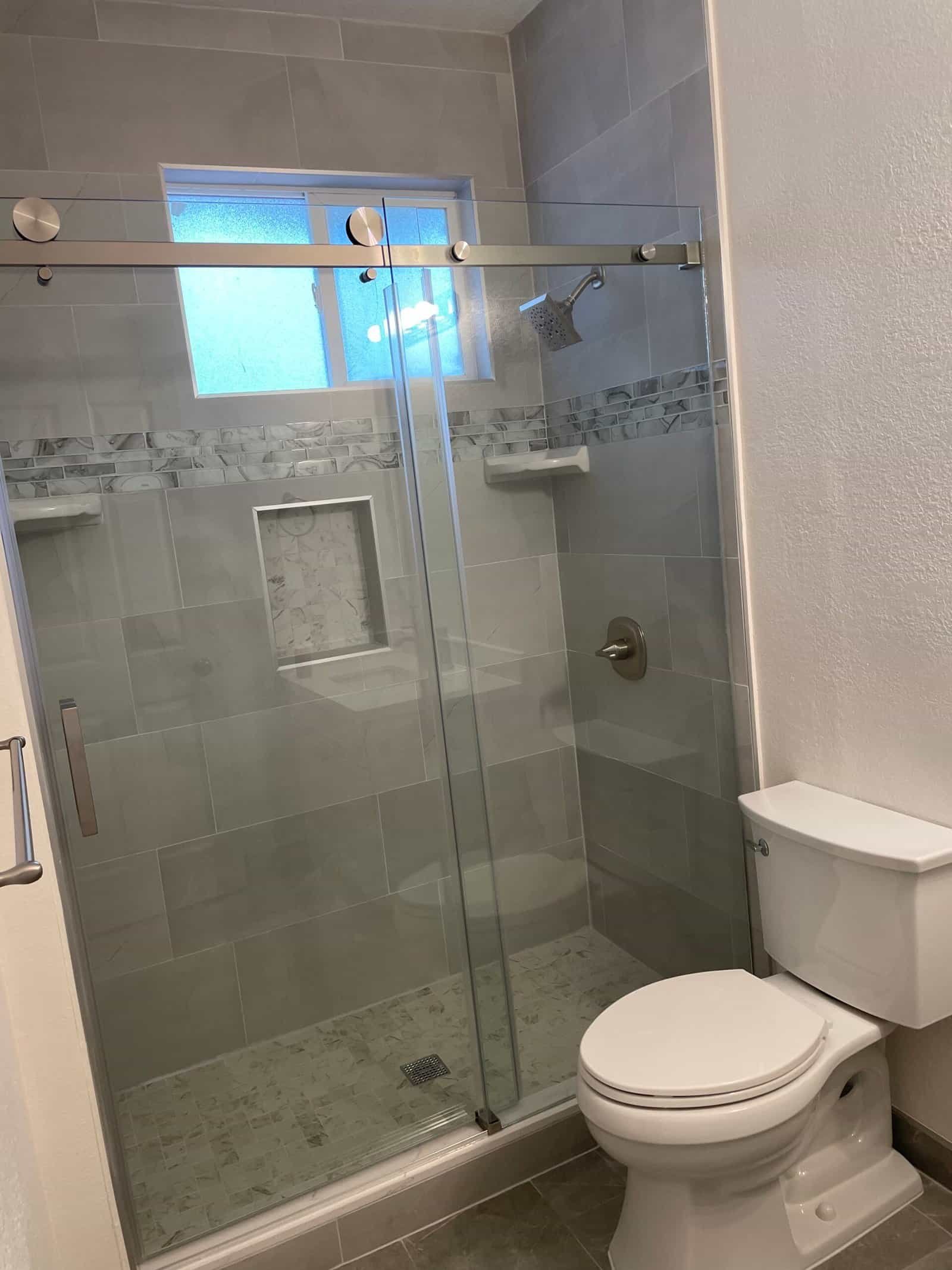 Full view of a contemporary bathroom shower with glass sliding doors, gray tiled walls, built-in niche, and a window for natural light.